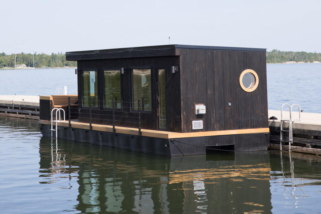 A modern black houseboat with large windows and a round porthole is docked at a pier on calm water.