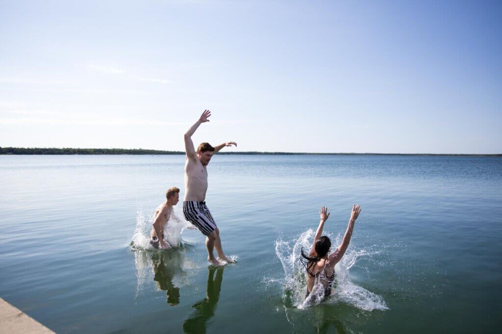 Three people splash and play in a calm lake on a sunny day, with two jumping into the water and one standing nearby.